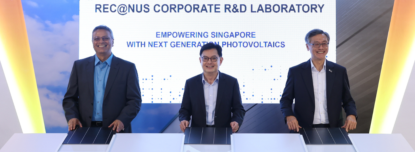 Three men in suits holding REC solar panels stand before a screen reading "REC@NUS Corporate R&D Laboratory."